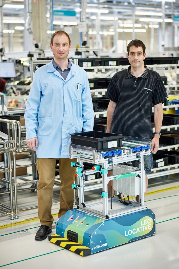 Lean Consultant Stepan und Pavel, Testsupport im Produktbereich elektromagnetische Sensoren (EMS) Pavel bei Pepperl+Fuchs in Tschechien, stehen in der Produktionshalle und lächeln in die Kamera. Vor ihnen steht das LEO-Locative-Transportsystem.