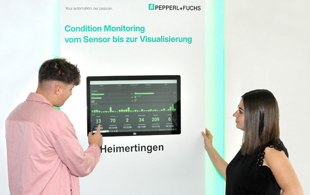 Man sieht das Dashboard in der Mitte des Bildes. Auf dem Display werden die Füllstände der smarten Kaffeemaschine angezeigt. Links vor dem Display steht ein Mann, der erklärend mit deiner rechten Hand auf einen der Werte zeigt. Rechts vor dem Display steht eine Frau, die interessiert auf das Display schaut und dem Mann zuhört.