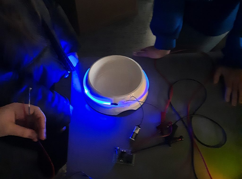 Das Bild zeigt den Hundenapf in seiner Entwicklungsphase. Er steht auf einem Tisch im Dunkeln, hat einen blau leuchtenden Beleuchtungsring um sich, von dem einige Kabeln abgehen. Auf dem Tisch sieht man zudem einen Mikrocontoller liegen.