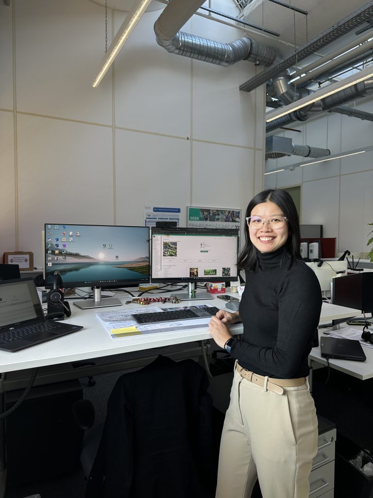 Yun Xia steht an ihrem Arbeitsplatz bei Pepperl+Fuchs und schaut in die Kamera. Auf ihrem Schreibtisch stehen ein Laptop, zwei Bildschirme und verschiedene Büromaterialien.