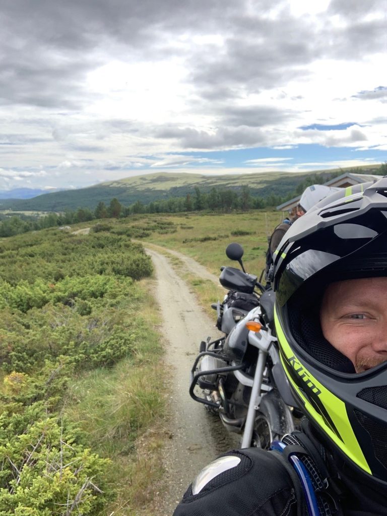Robert steht auf einem schmalen, unbefestigten Weg, der sich durch eine grüne, hügelige Landschaft schlängelt. Es ist bewölkt. Er steht vor seinem Motorrad und macht ein Selfie von sich. Er trägt Motorradkleidung und lächelt in die Kamera.