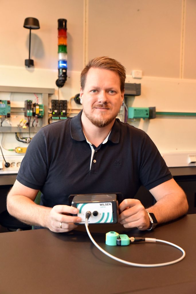 Robert, System & Solution Engineer, an seinem Arbeitsplatz. Er sitzt am Tisch und hat vor sich den Wilsen-Sensor in der Hand. Hinter ihm sind verschiedene Geräte zu sehen.