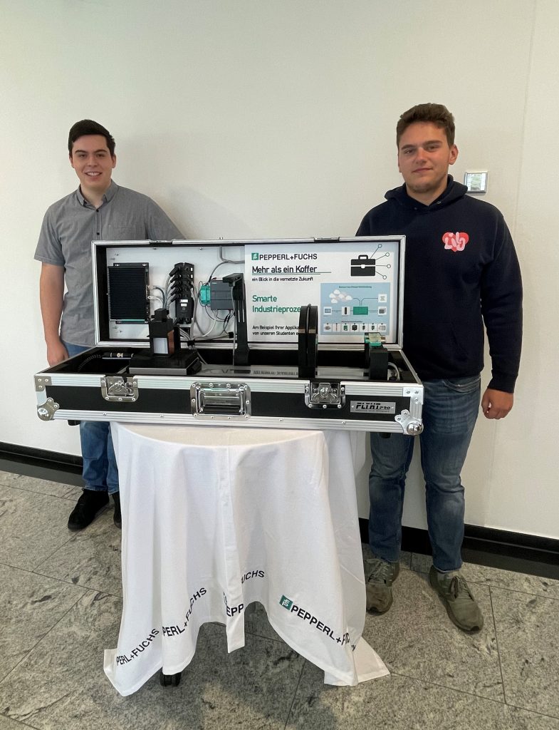 Praxisphasen bei Pepperl+Fuchs: Dennis und Aaron stehen hinter dem geöffneten Kofferexponat, welches auf einem runden Tisch mit einem weißen Tischtuch mit Pepperl+Fuchs Logo steht. Im Koffer sieht man die verbaute Sensorik.
