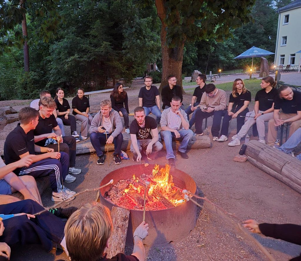 Die dual Studierenden und Auszubildenden sitzen um eine Feuerschale herum. Es dämmert bereits, mache von ihnen halten an einem Stock Marshmallows ins Feuer.
