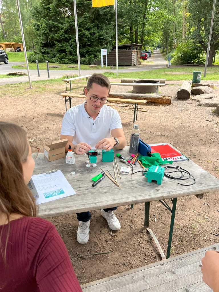 Ein junger Mann sitzt draußen an einem Tisch. Am Rand sind noch zwei weitere Personen zu erkennen. Auf dem Tisch liegen verschiedene Materialien, sowie verschiedene Technik/Sensorik. Er bastelt daran herum.