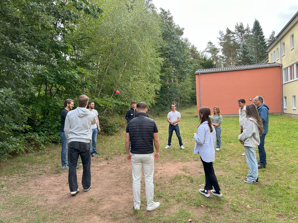 Eine Gruppe von elf Personen steht in einem Kreis auf einer Wiese und wirft sich gegenseitig einen Ball zu. Im Hintergrund ist die Jugendherberge zu sehen.