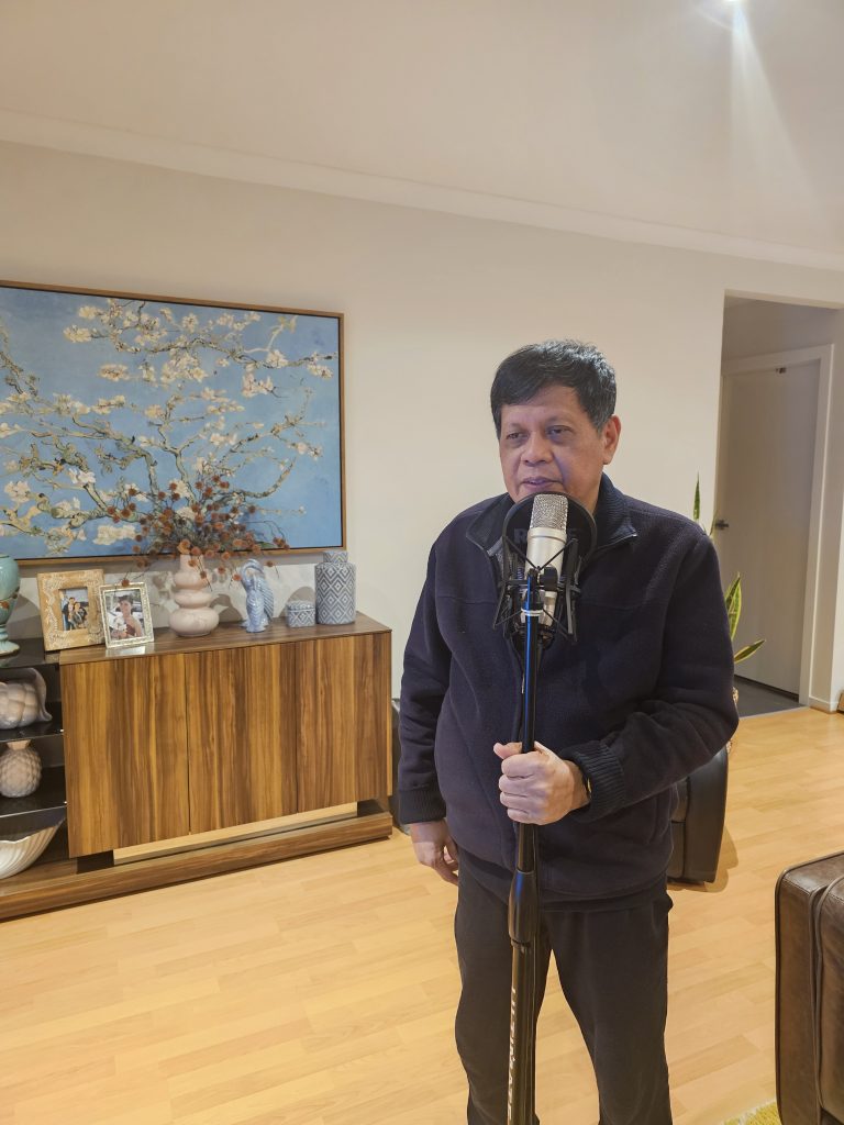 Benjamin steht bei sich zuhause im Wohnzimmer und singt in ein Mikrofon. Hinter ihm ist ein Sideboard mit privaten Bildern zu sehen, sowie ein großes Gemälde an der Wand.