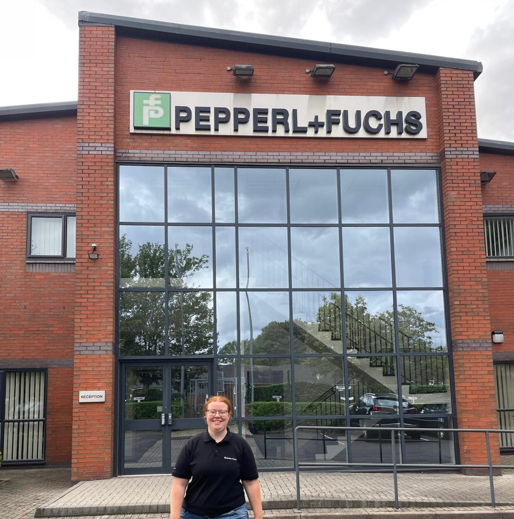 Melanie absolviert ein Auslandssemester in Oldham. Hier steht sie vor dem Eingang des Pepperl+Fuchs Gebäudes in Oldham. Es ist ein rotes Backsteingebäude mit einer großen Glasfront direkt hinter ihr.
